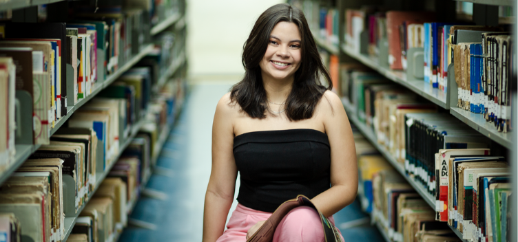 #ParaTodosVerem:Fotografia colorida mostra jovem sorrindo entre duas prateleiras de livros. Ela está sentada e tem uma revista apoiada sobre a perna esquerda. A jovem tem cabelos lisos e pretos, na altura dos ombros. Veste uma blusa preta, modelo tomara que caia, e calça comprida na cor rosa.