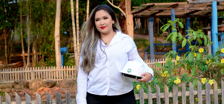 #ParaTodosVerem: Fotografia colorida de uma jovem de cabelo liso, longo e claro. Ela está de pé, usa batom vermelho, colar e brincos dourados e veste uma camisa de mangas compridas na cor branca. Ela está em um ambiente natural com presença de muitas árvores e carrega um capacete de segurança na mão esquerda. 