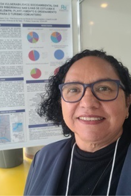 #ParaTodosVerem: Fotografia colorida, em formato 3x4, mostra uma mulher de cabelo curto, preto e encaracolado. Ela veste blazer de cor azul, com blusa preta e usa óculos. Ela sorri e está em pé à frente de um banner de pesquisa.