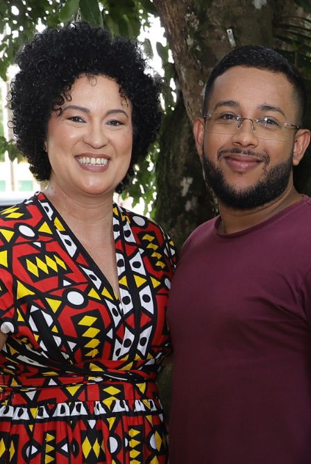 #ParaTodosVerem:Fotografia mostra um homem e uma mulher, um ao lado do outro. A mulher tem cabelos pretos e curtos. Ela traja roupa com estampa étnica e colorida e sorri. O homem tem cabelos curtos. Ele usa barba e bigode, está de óculos e veste uma blusa de cor roxa. 