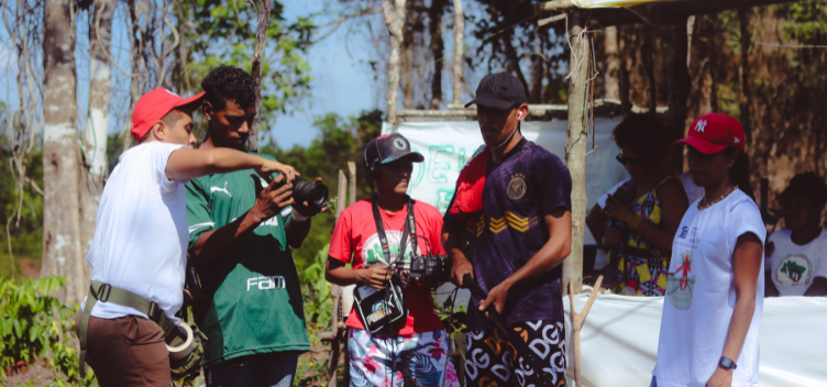 #ParaTodosVerem: Fotografia mostra, em primeiro plano, cinco pessoas. Três delas seguram equipamentos de fotografia e filmagem. À direita, outras pessoas observam a cena, em uma barraca, protegidas do sol. Ao fundo, veem-se árvores de diversas espécies.