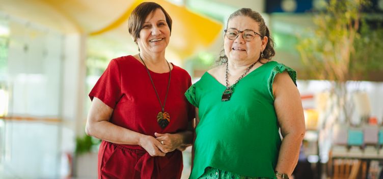 #ParaTodosVerem: Fotografia mostra duas mulheres em pé diante de um salão. A da esquerda tem cabelos curtos e castanhos. Ela usa brincos e um colar com pingente em formato de folha em tons verde e laranja. Está vestida com uma calça e blusa de cor vermelha. A da direita tem cabelos castanhos com mechas brancas acima do ombro. Ela usa óculos, um par de brincos e um colar de correntes com uma pedra preta como pingente. Está vestida com uma blusa e saia na cor verde.