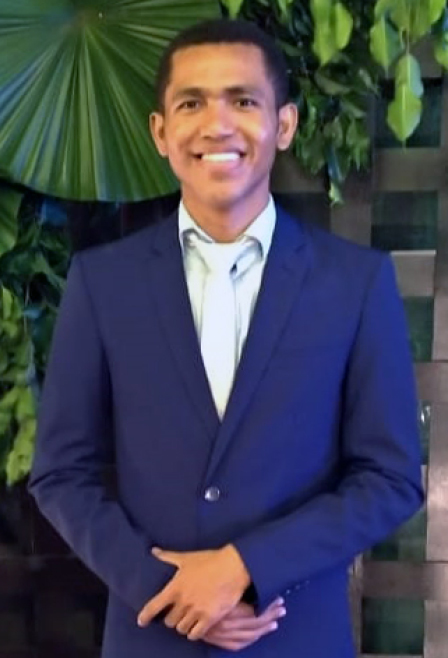 Na fotografia, homem jovem, negro, com cabelos e olhos escuros, veste terno azul-marinho. No fundo, um painel decorado com folhagem natural. O homem olha e sorri para nós.