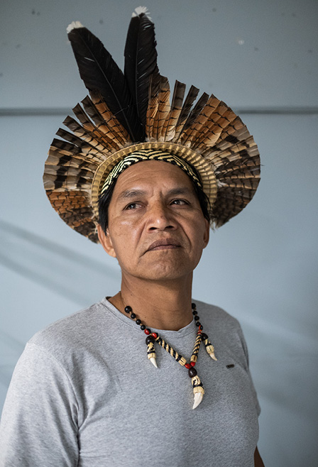 #ParaTodosVerem: Fotografia com fundo branco mostra um homem indígena. Ele usa cocar e colar, veste uma camiseta cinza de mangas curtas e direciona o olhar para a esquerda.