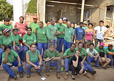 #ParaTodosVerem: Fotografia mostra grupo com mais de 20 pessoas, homens e mulheres organizados, sentados e em pé. Todos olham em nossa direção. Alguns vestem uma espécie de uniforme composto por calça azul e blusa verde. Ao fundo, material reciclável à espera da triagem.