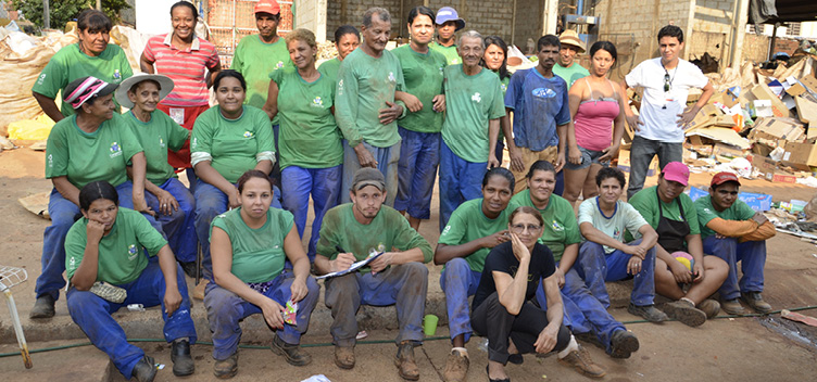 #ParaTodosVerem: Fotografia mostra grupo com mais de 20 pessoas, homens e mulheres organizados, sentados e em pé. Todos olham em nossa direção. Alguns vestem uma espécie de uniforme composto por calça azul e blusa verde. Ao fundo, material reciclável à espera da triagem.